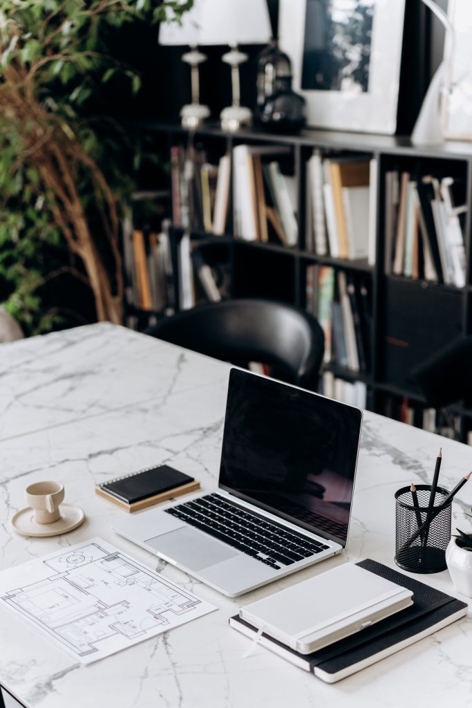 Laptop, closed notebook, coffee cup, and floor plan drawings on a marble table.