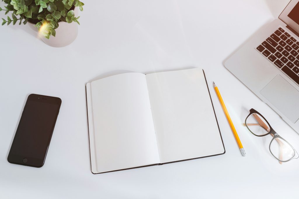 Open blank notebook beside a smartphone, laptop, glasses, and pencil on a white desk.